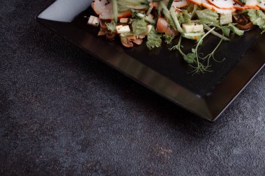 Summer salad with pieces of bacon, fresh cucumbers, cheese cubes and mushrooms. Green salad on a black plate on a dark background. Free space for an inscription. View from above.