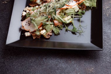 Summer salad with pieces of bacon, fresh cucumbers, cheese cubes and mushrooms. Green salad on a black plate on a dark background. Free space for an inscription. View from above.
