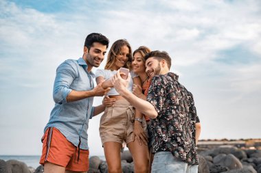 Bir grup genç arkadaş deniz kenarında yan yana duran akıllı telefondan sosyal medya içeriklerine bakıyorlar. Cep telefonu kullanan Z jenerasyonunun alt görüntüsü.