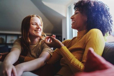 Çok kültürlü eşcinsel kadınlar kanepede oturmuş neşeli konuşuyorlar. Lezbiyen çift evde eğleniyor.