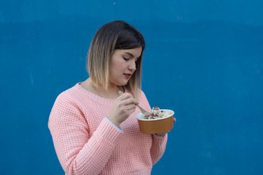 Açık havada salata yiyen sevimli kadın mavi arka planda izole edilmiş plastik organik çatal bıçak kullanıyor.