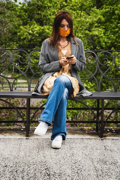 A 20 year old young woman wearing a fashion face mask sitting outdoors using smartphone alone - one girl with crossed legs sitting on a bench in the park typing on smartphone screen