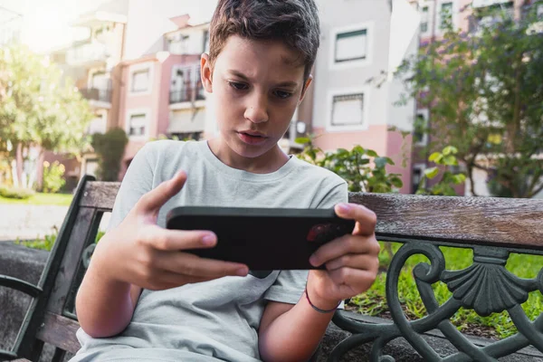 屋外のベンチに座ってソーシャルネットワークコンテンツをオンラインで探しているデジタルデバイスを使用して小さな男の子 — ストック写真