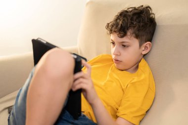 Okul çağındaki bir çocuk kapalı alanda koçu rahatlatan bir tablet kullanıyor. Teknolojiden bağımlı çocuklar evden eğitim, bağımlılık, insanlar ve teknoloji konsepti