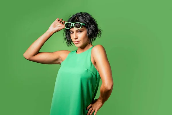 Winking young brunette woman 20s years old wears tank top touching raise sunglasses up - isolated on plain vivid green background - studio portrait - People emotions lifestyle concept