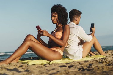 Yazın plajda omuz omuza otururken birden fazla etnik çeşitliliğe sahip genç çiftler, her zaman internete bağlı gençler, teknoloji ve insan yaşam tarzı kavramı.