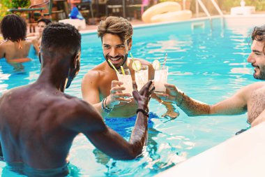 Multiethnic group of young friends enjoying a swimming pool party - young adult men clinking toasting glasses of mojito cocktails and having fun together - people, food and drink lifestyle concept