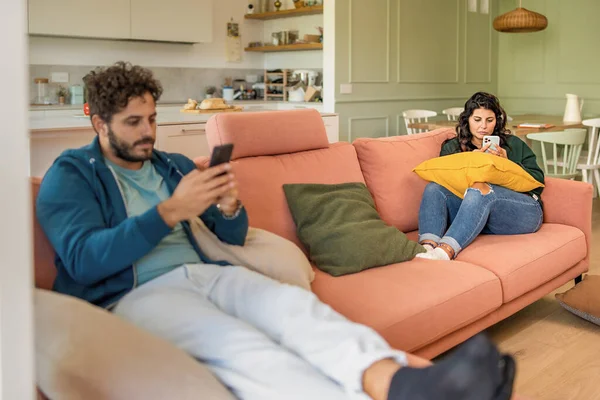 Young married couple using smart phone ignoring each other addicted to the internet and social network - man and woman phubbing in the living room - family people loneliness and technology lifestyle concept