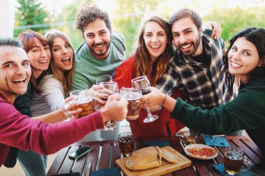 Group of cheerful young friends cheering with wine and beer glasses at picnic happy hour party in the terrace - Young people having fun drinking and eating outdoor - Friendship and youth holidays lifestyle concept