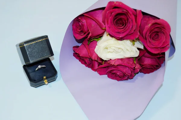 蓝色的背景上摆放着一束紫色包装的红玫瑰和一个有戒指的盒子 婚姻建议的概念 — 图库照片