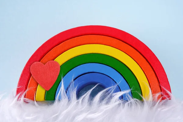 Wooden toy rainbow with a heart on a blue background with white fur, similar to a cloud below.Montessori concept, autism, environmental toys, art therapy, kindergarten, playroom, rainbow party, LGBT community