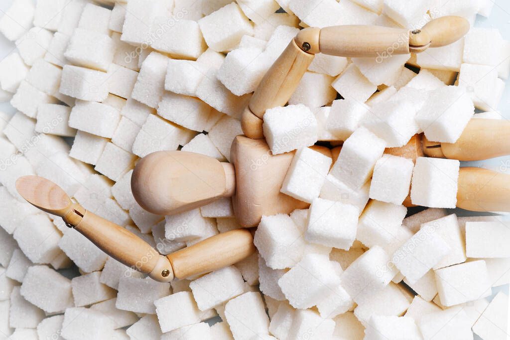 A wooden mannequin doll, strewn with sugar cubes, is drowning in a pile of sugar.Unhealthy concept of sugar addiction, obesity and related diseases, diabetes, articles about the dangers of sugar.