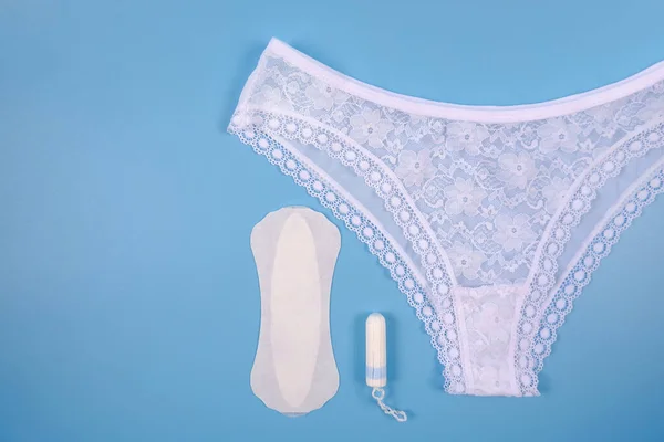 Beyaz seksi dantel külot, günlük hijyenik pedler, mavi arka planda tamponlar. Kritik günlerde giysi konsepti, regl döneminde temizlik, kadın hijyeni ve sağlık.