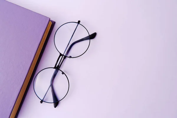 A blue-purple book or notebook (the fashionable color of 2022) with round stylish optical glasses on a lilac background.The concept of reading books, planning, making a to-do list, libraries.Copyspace