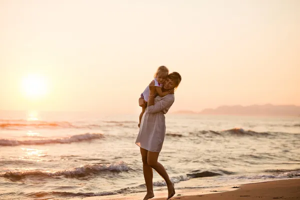 Glad mamma håller sin dotter på stranden vid solnedgången, sommar — Stockfoto