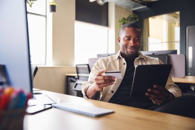 Smiling Man making online payment on tablet computer holding credit card in office