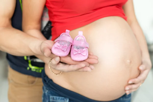 Foto de una mujer atractiva que está embarazada de 38 semanas sosteniendo botines de bebé rosados y blancos en su barriga . — Foto de Stock
