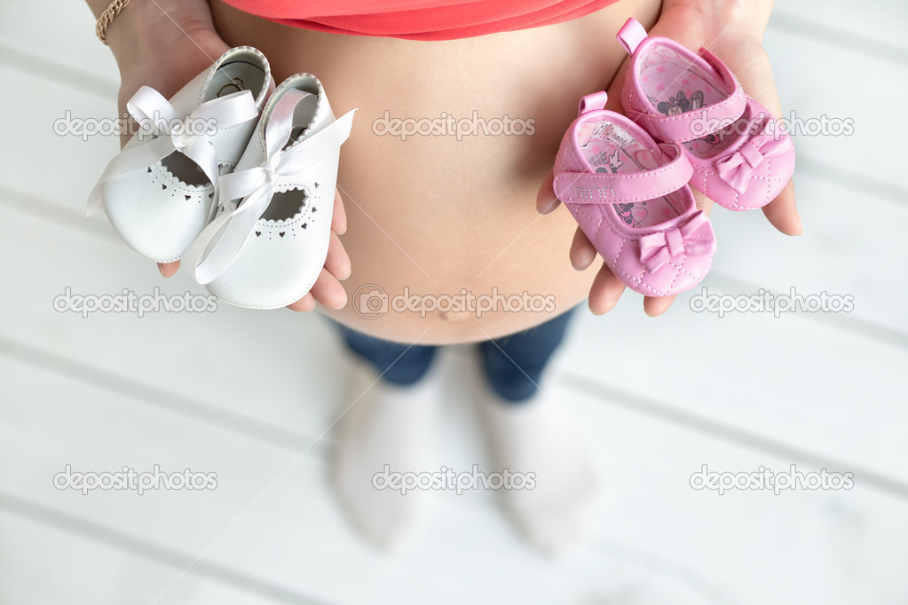 Photo of an attractive woman who is 38 weeks pregnant holding pink and white baby booties on her tummy.