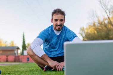 Çevrimiçi bir sınıfta spor giyinen sağlıklı bir adam bilgisayarını monitörü ve kişisel antrenörüyle açık havada antrenman yapmayı öğreniyor.