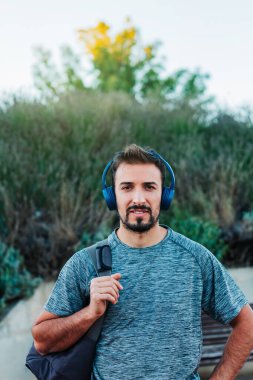 Dikey görüntü. Spor kıyafetleri ve çanta giymiş genç bir adam mavi kulaklıklarıyla sokakta yürüyor müzik dinliyor ve kameraya bakıyor.