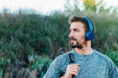Spor kıyafetleri ve çanta giymiş genç bir adam spor salonuna giderken yolda yürürken mavi kulaklıklarıyla kedileri dinliyor..