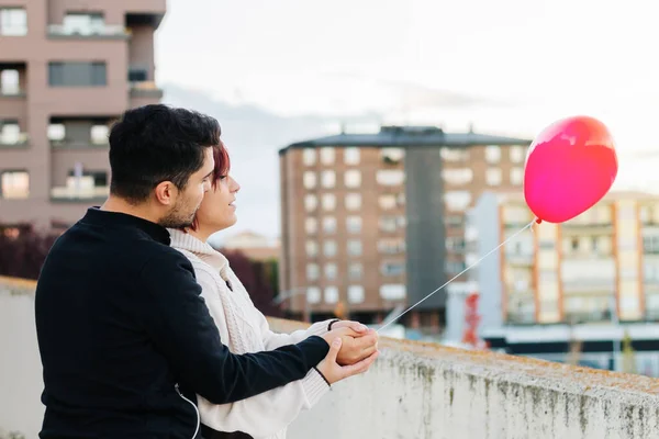 Young couple, boy and girl, with their backs turned and unrecognizable, holding a red heart-shaped balloon on a balcony with the city in the background. Copy space, love concept, valentine.