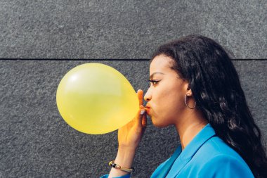 Siyah saçlı, sarı balonu şişiren, gri arka planda duran Afro-Amerikan kadın.
