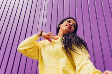 Mor arka planda sarı elbiseli şehir dansçısının alt görüntüsü. Açık havada dans eden genç bir kadının düşük açılı çekimi, şehir tarzı ve gençlik moda konsepti.