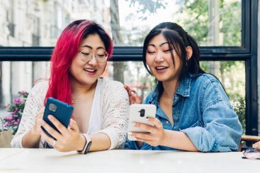 Asyalı genç bayan arkadaşlar, mutlu konuşuyorlar ve gülümsüyorlar, cep telefonlarına bakıyorlar. Genç Japon kız kardeşler barda akıllı telefonlarına rahatça bakıyorlar..