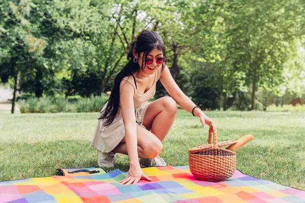 Young Asian girl putting down the picnic blanket at a gathering with her friends to eat outdoors. Attractive Japanese woman with a picnic basket enjoying a summer day outdoors on vacation.