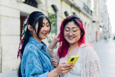 Günlük kıyafetlerle gezen genç bir Japon kadın sosyal medyada cep telefonuyla mutlu kız kardeşiyle video paylaşırken gündüz vakti şehrin bulanık arka planında dikiliyor.