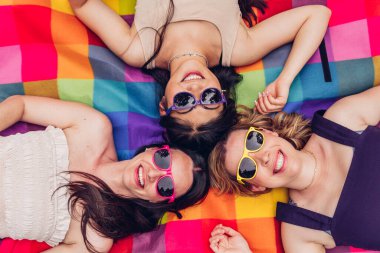 Moda gözlüklü, neşeli, çok kültürlü kadınların hafta sonu pikniği sırasında renkli kareli battaniyenin üzerinde dinlenirken gülümseyip kameraya baktığı en iyi manzara.