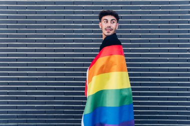 Yüzünde renkli gökkuşağı LGBT bayrağı olan homoseksüel bir adam omzunun üstünden bakarken sokakta duvarın yanında dikiliyor.