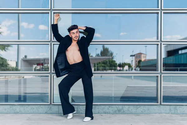 Full body of queer male in black jacket on naked torso standing with raised arm near glass wall in city