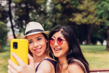 Güneşli bir günde yeşil parkta dikilirken akıllı telefondan selfie çeken gündelik kıyafetli genç kızlar.