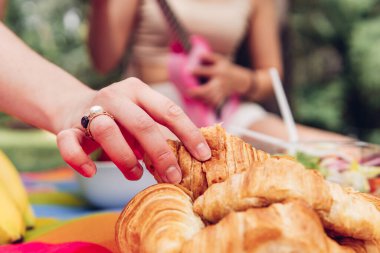 Close-up of an unrecognizable woman's hand picking up a croissant at a picnic with friends. Anonymous person having french butter croissant breakfast outdoors in the morning
