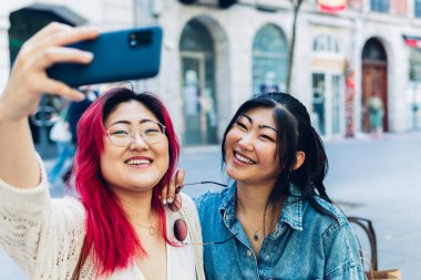 İki güzel Asyalı kadın, büyük bir gülümsemeyle akıllı telefonlarıyla selfie çekiyorlar..