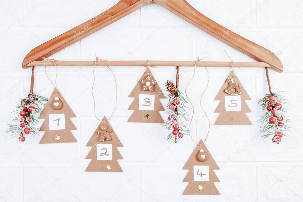 Craft Christmas trees with numbers 1,2,3,4,5 and fir branches with red berries hang on a wooden hanger on a white brick wall, close-up side view. Advent calendar concept.