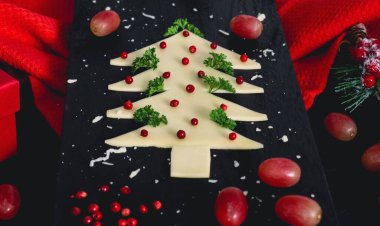 Kırmızı bir mutfak peçetesiyle kesim tahtasındaki peynirden, baharatlardan ve üzümlerden oluşan yaratıcı Noel ağacı siyah arka planda, yakın planda yatar..