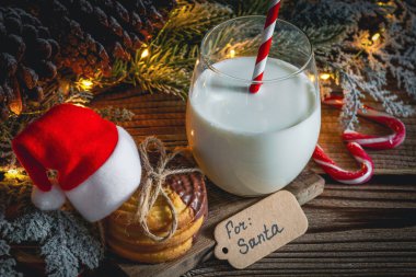 Tahtada süt ve Noel Baba şapkası olan kurabiyeler, Noel ağacı, çam kozalakları, karamel şekerler ve üzerinde şöyle yazan bir not: Masanın üstünde Noel Baba için, yakın çekim..