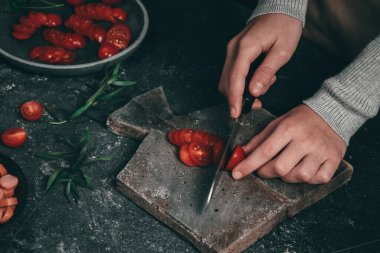 Genç bir kadının elleri, koyu arka planda, yakın planda gri bir tahta üzerinde büyük siyah bıçaklı vişneli domateslerle kesiyor. Yemek pişirme konsepti.