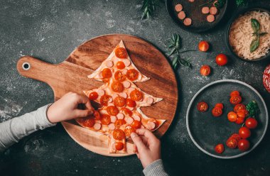 Çocuklar dilimlenmiş kiraz domateslerini pizza hamurundan kesilmiş bir Noel ağacının üzerine koyuyorlar. Kesme tahtası üzerinde koyu bir zemin üzerinde uzanıyorlar, mozzarella peyniri, sosis ve dekorasyon, yakın çekim. Noel ve Yeni Yıl için hazırlık konsepti