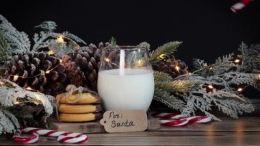 Bir el, bir bardağa bir pipet koyar ve masanın üzerinde kurabiyeler, bir ladin dalı, koniler, şekerler, bir çelenk ve bir not vardır: Noel Baba için ahşap bir masa, yakın çekim kenar görünümü..