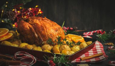 Patatesli, otlu, portakallı ve tarçınlı tavuk. Mutfakta kırmızı kareli bir peçete ve koyu arkaplanda bir noel süsü..