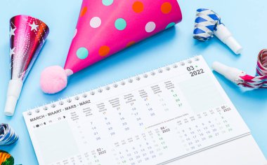 Doğum günü setiyle birlikte Mart 2022 için masaüstü kağıt takvimi: ıslıklar, borular, kağıt koni şapkalar, açık mavi bir arkaplanda uzanan renkli süsler, yakın çekim yan görünümü. Takvim, doğum günü ve planlama kavramı.