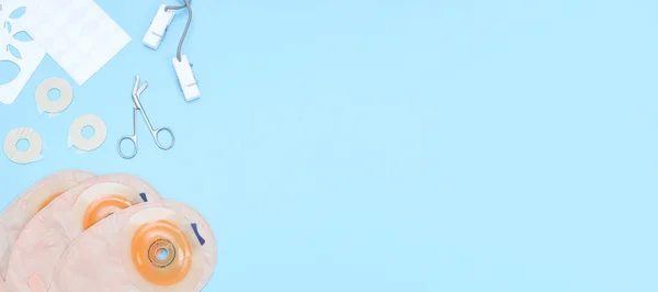 Three colostomy bags with medical scissors, a stencil of different diameters, a sticky circle and clothespins on a rope lie on the left on a soft blue background with copy space on the right, flat lay close-up.