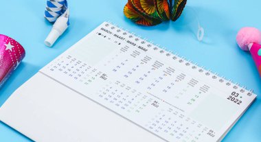 Doğum günü setiyle birlikte Mart 2022 için masaüstü kağıt takvimi: ıslıklar, borular, kağıt koni şapkalar, açık mavi bir arkaplanda uzanan renkli süsler, yakın çekim yan görünümü. Takvim, doğum günü ve planlama kavramı.