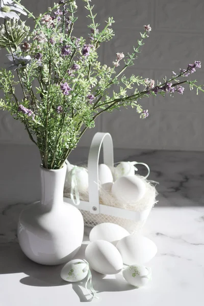 Decoratieve witte eieren, metalen mandje met eieren en een vaas met lentebloemen op een marmeren tafel. — Stockfoto