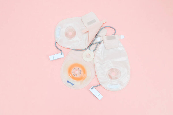 Three colostomy bags with medical clips lie on a pale pink background,