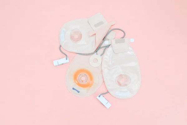 Three colostomy bags with medical clips lie on a pale pink background,